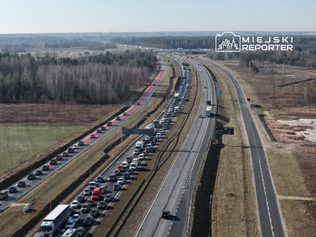 Zatłoczona droga ekspresowa z wieloma pojazdami stojącymi w korku, w otoczeniu drzew i pustych pól.