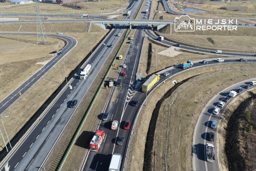 Ruch samochodowy na skrzyżowaniu dróg, z pojazdami na różnych pasach ruchu oraz widoczną infrastrukturą drogową.
