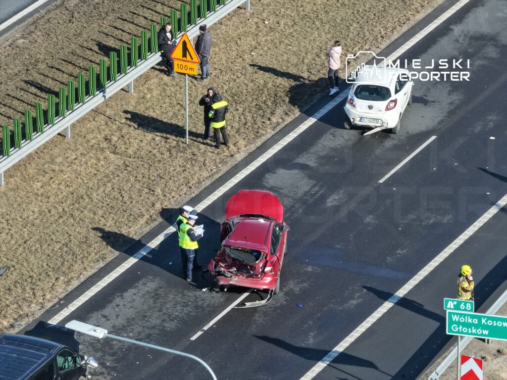 Policjant w żółtej kamizelce dokonuje oględzin zderzonego czerwonego samochodu na autostradzie, obok stoi uszkodzony biały pojazd.