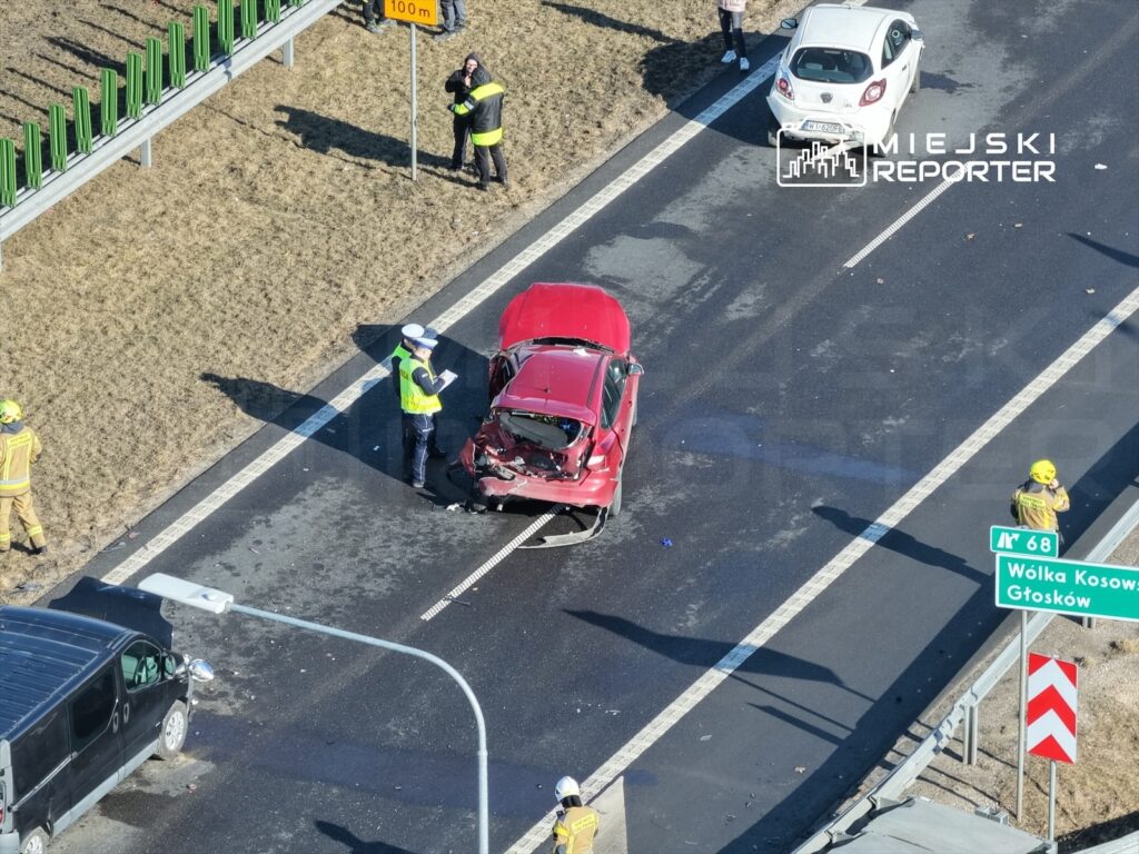 Czerwony samochód osobowy z uszkodzeniami stoi na jezdni, obok pracują strażacy i policjant spisujący zdarzenie.