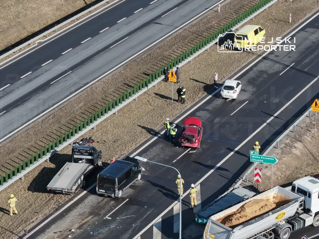 Na drodze widać wypadek samochodowy z uszkodzonymi pojazdami, a obok pracują strażacy i policjanci.