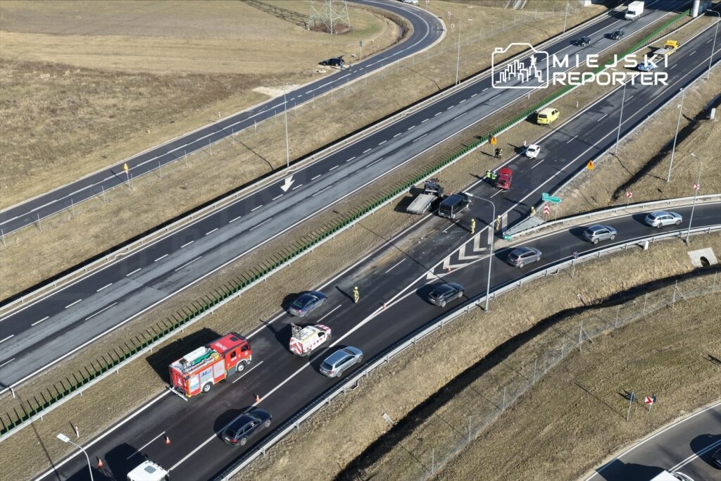 Wielu ratowników i pojazdów na drodze po wypadku, z uszkodzonymi samochodami na pasie ruchu.