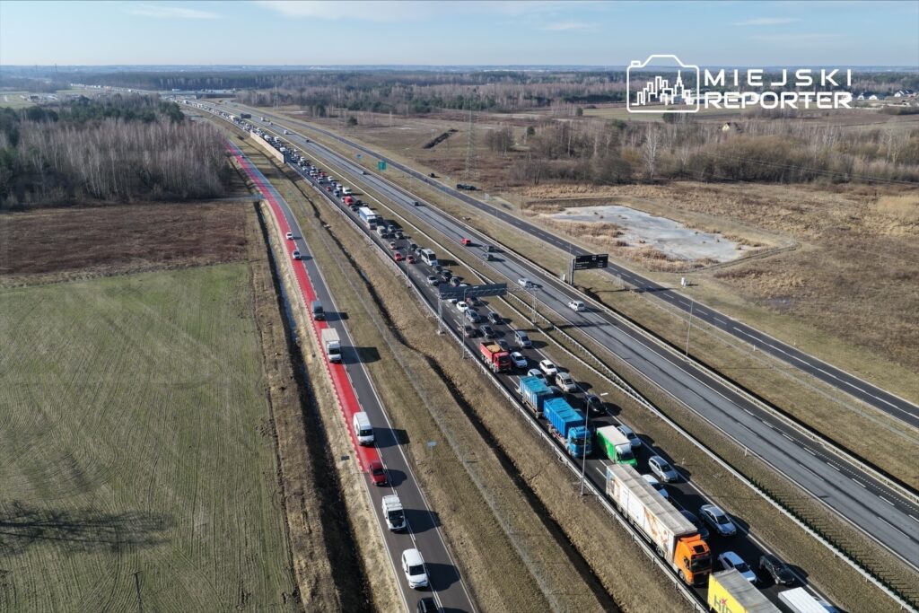 Zatłoczona droga ekspresowa z samochodami osobowymi i ciężarówkami w kierunku przeciwnym, otoczona polami.