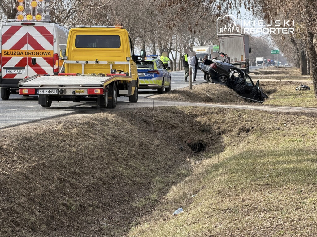 Na poboczu drogi stoi żółta laweta, obok niej policyjny radiowóz oraz wóz służby drogowej, a w rowie leży przewrócone auto.