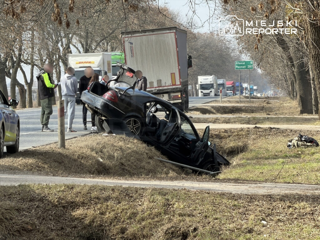 Czarny samochód osobowy w rowie, obok grupka osób rozmawiających, w tle ruch drogowy z ciężarówkami.