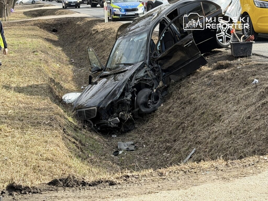 Czarny samochód osobowy w rowie, obok drogi, z uszkodzoną przednią częścią i otwartymi drzwiami.
