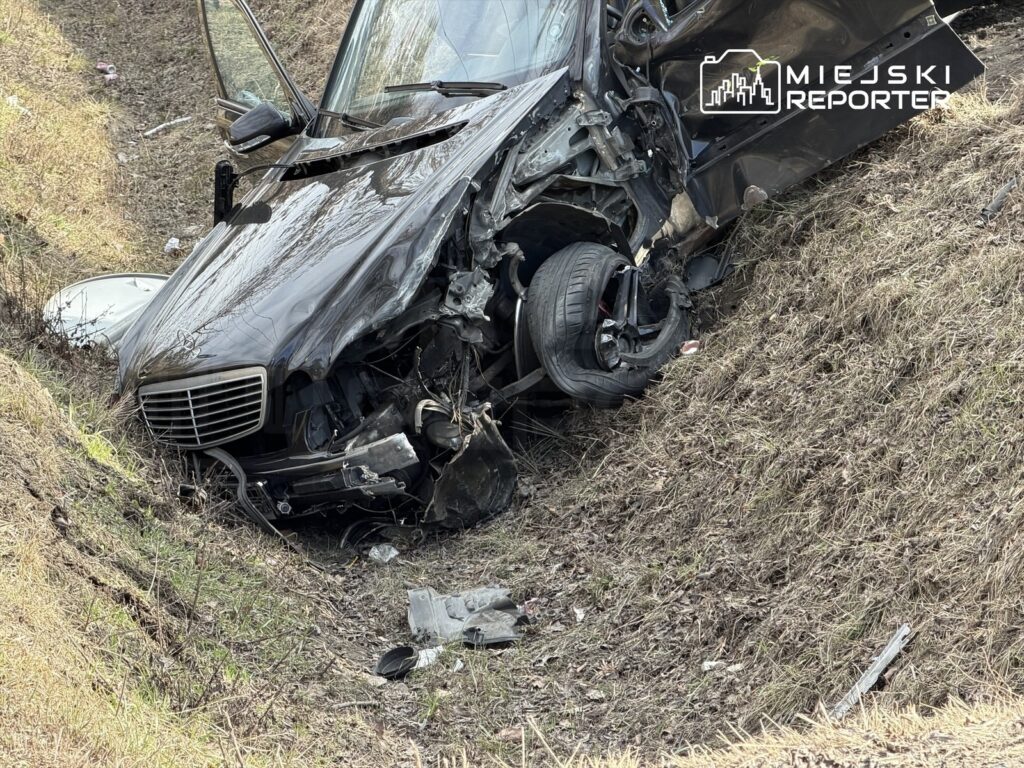 Czarny samochód osobowy leży na poboczu drogi, z uszkodzoną przednią częścią i jednym kołem uniesionym w powietrzu.