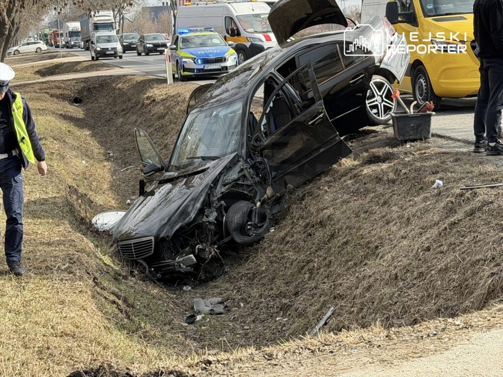 Czarny samochód osobowy w rowie, obok stoi policjant w kamizelce odblaskowej, na tle ruchliwej drogi.