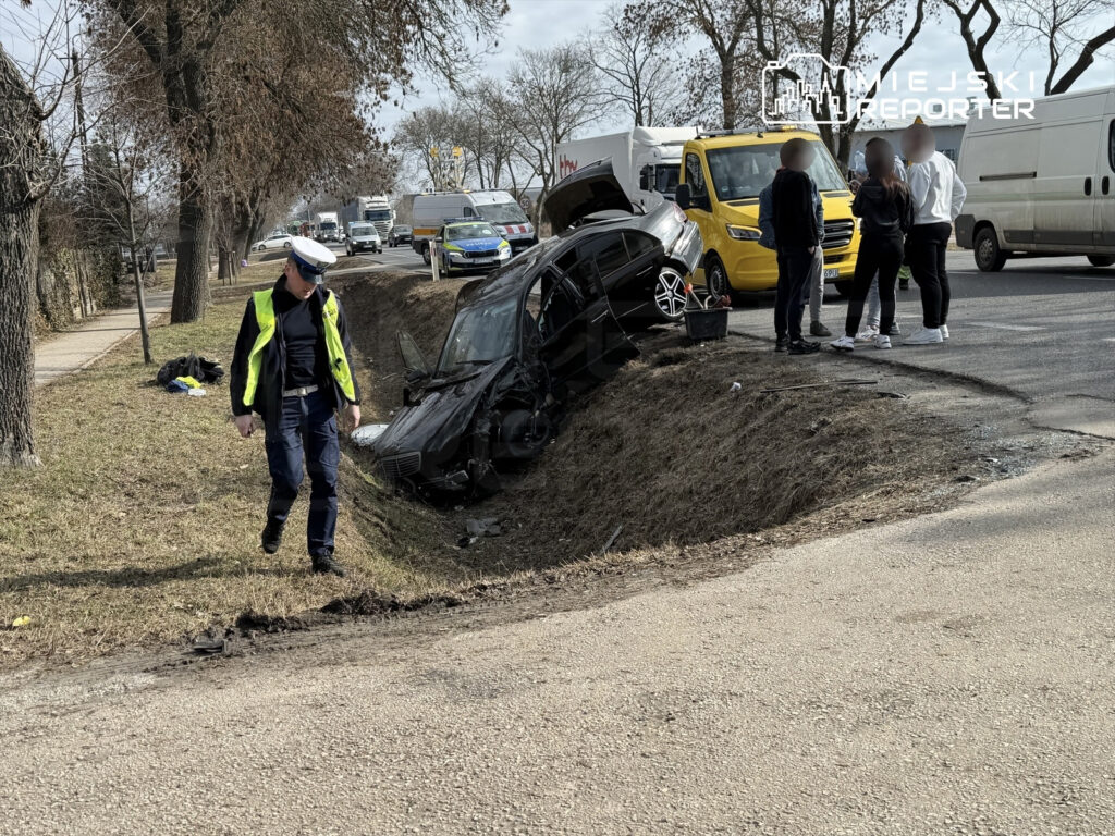 Policjant w odblaskowej kamizelce bada miejsce wypadku, obok stoi grupa osób, a w rowie leży uszkodzony samochód.