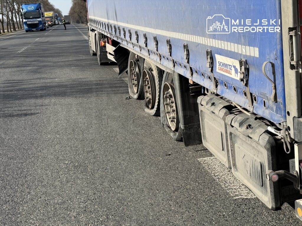 Ciągnik siodłowy z uszkodzoną oponą zatrzymany na drodze, w tle widoczne inne pojazdy.