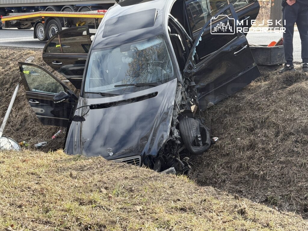Czarny samochód osobowy w rowie, z otwartymi drzwiami, obok leżący słup drogowy oraz osoba w pobliżu.
