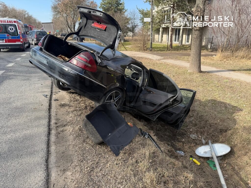 Czarny samochód osobowy leży na boku na poboczu drogi, z otwartymi drzwiami i bagażnikiem, obok widoczne ślady wypadku.