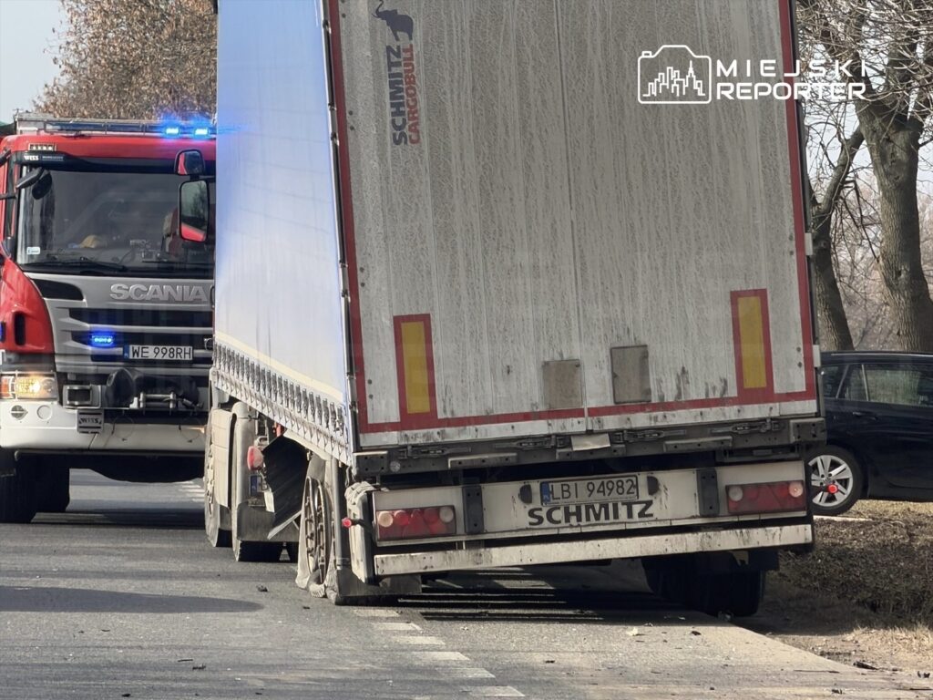 Czerwony wóz strażacki z włączonymi sygnałami świetlnymi stoi obok uszkodzonej ciężarówki z widocznym przebitym kołem.