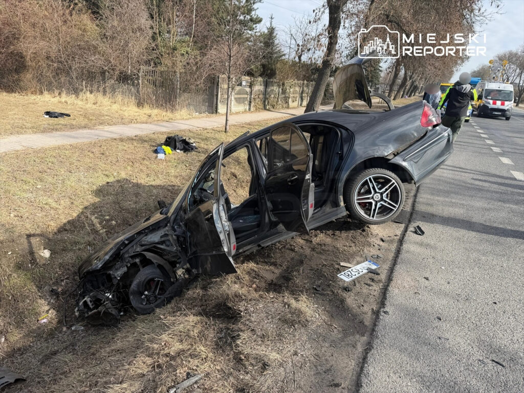 Czarny samochód osobowy leży na poboczu drogi, z otwartymi drzwiami, obok widać porozrzucane fragmenty pojazdu.