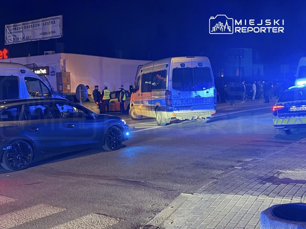 Grupa policjantów prowadzi działania na ulicy, obok zaparkowanych radiowozów i wozu strażackiego, w nocy.