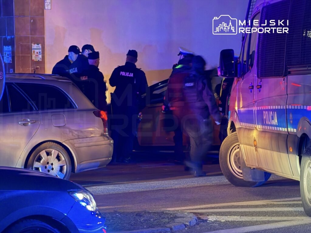 Grupa policjantów w mundurach prowadzi interwencję obok zaparkowanego samochodu w nocy.