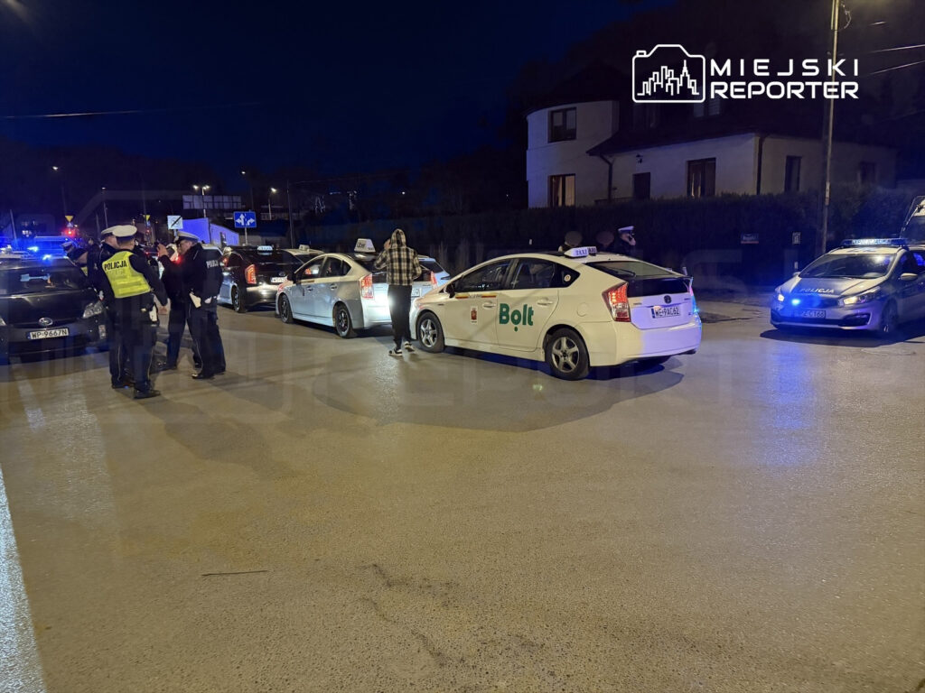 Grupa policjantów rozmawia z kierowcami taksówek na oświetlonej ulicy nocą.