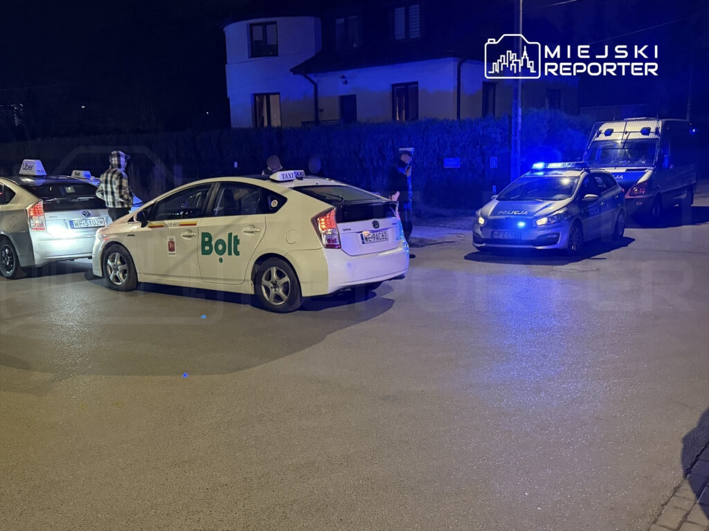 Taksówki z logo Bolt i Uber stoją na ulicy, obok nich policyjny radiowóz z włączonymi sygnałami świetlnymi.