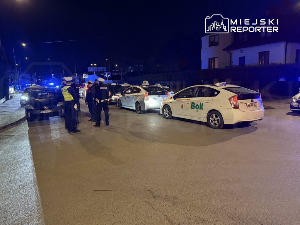 Grupa policjantów rozmawia na ulicy, obok zaparkowanych samochodów, w nocy z włączonymi światłami.