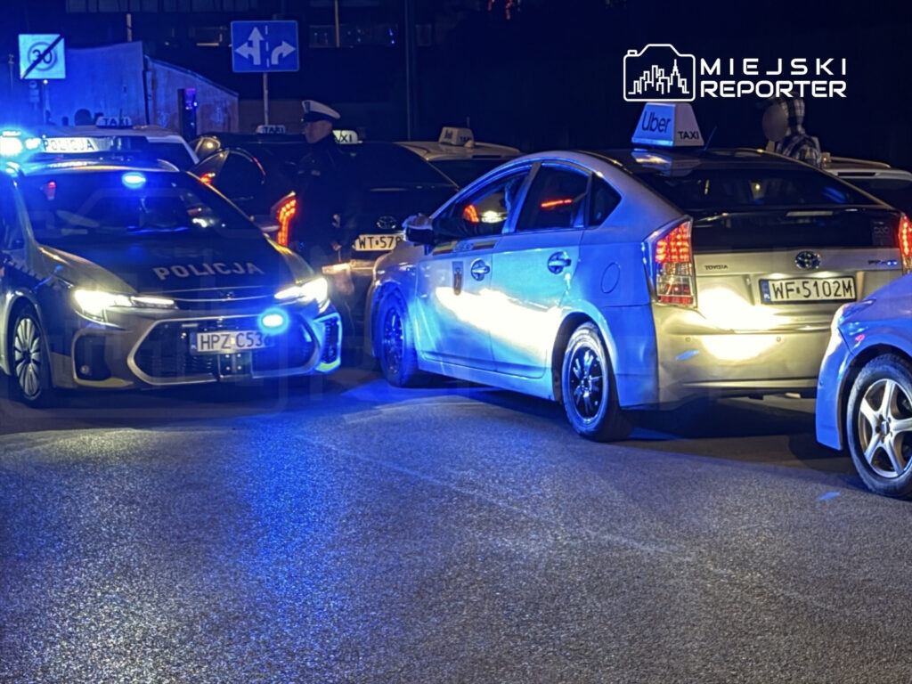 Policyjny radiowóz z włączonymi sygnałami świetlnymi zatrzymuje samochód osobowy na ulicy w nocy.