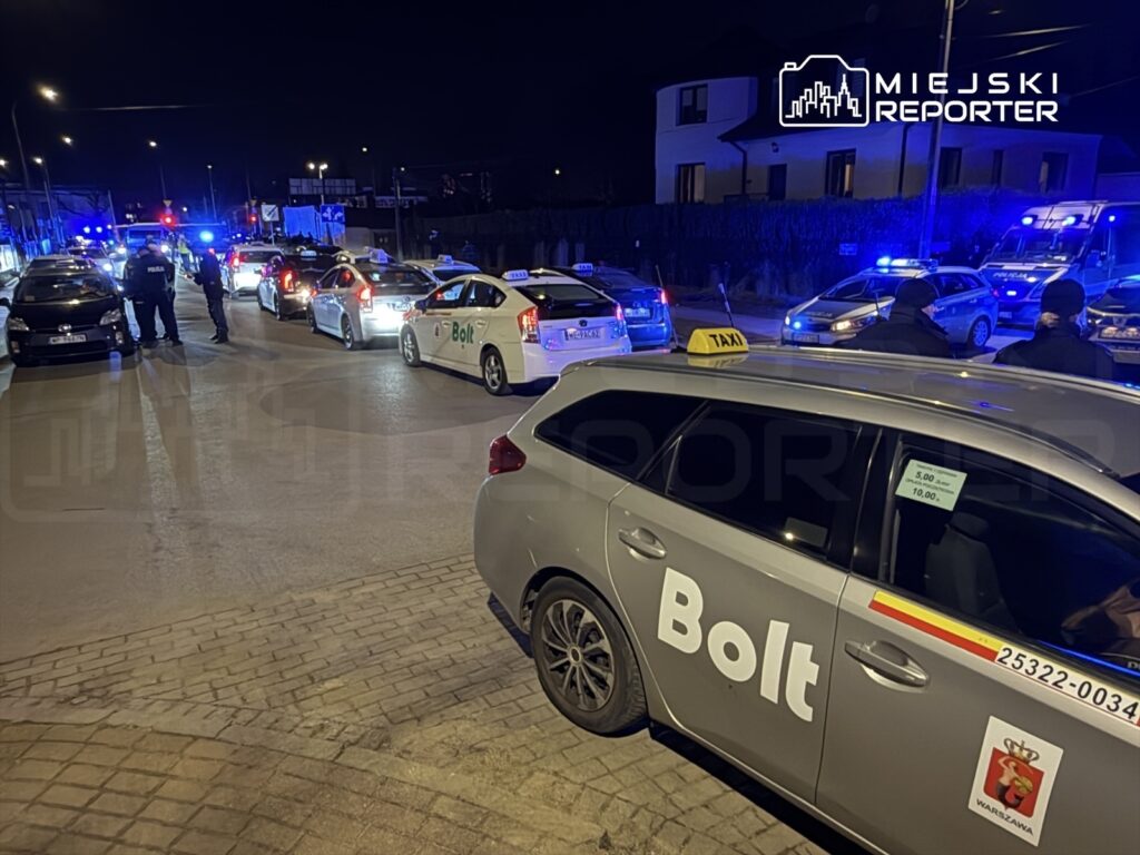Policyjni funkcjonariusze kontrolują taksówki na ulicy, w tle widać niebieskie światła radiowozów.