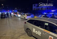 Policyjni funkcjonariusze kontrolują taksówki na ulicy, w tle widać niebieskie światła radiowozów.