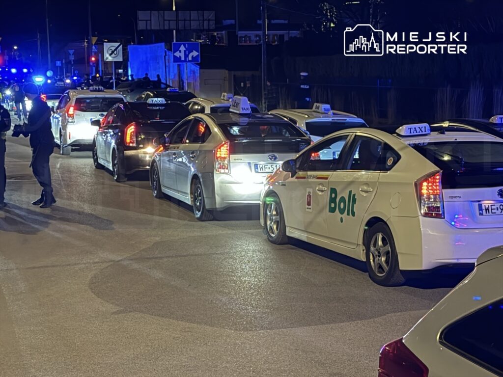 Rządowa kolumna taksówek i samochodów dostawczych zatrzymana na ulicy w nocy, z widocznymi światłami policyjnymi.