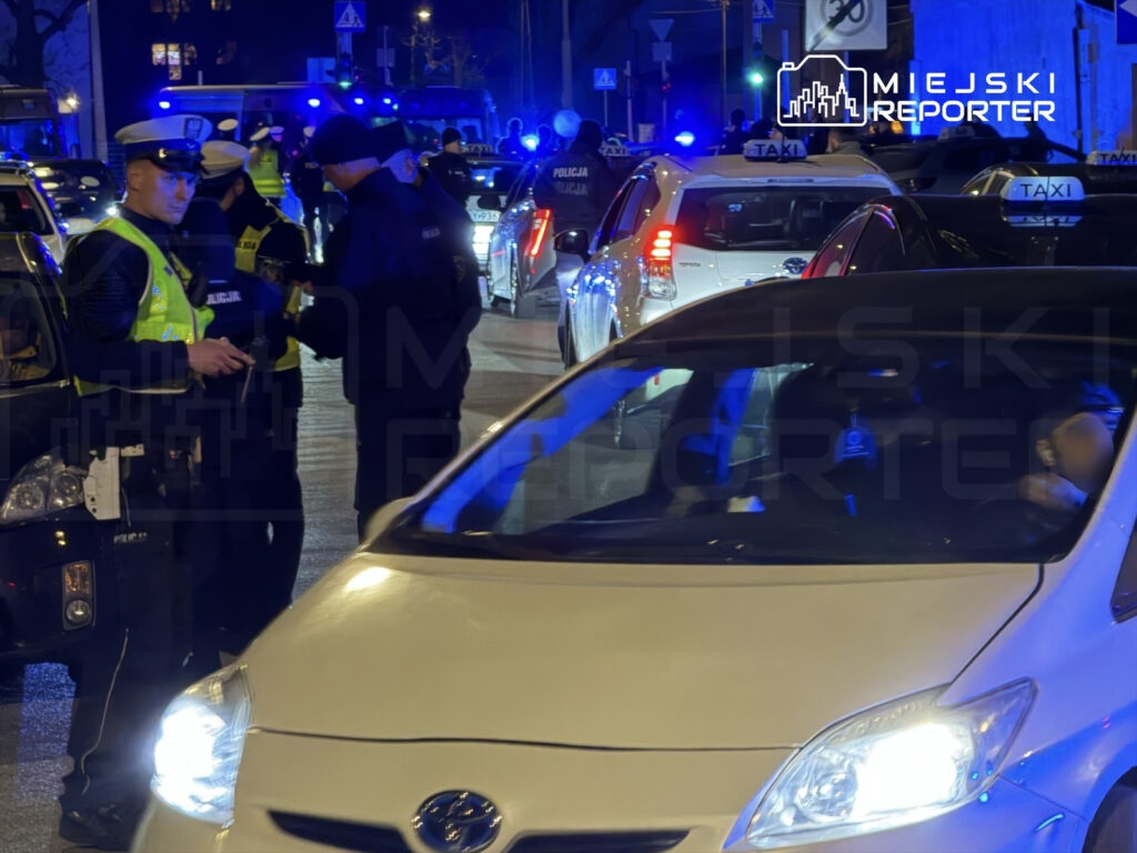 Policjanci w kamizelkach odblaskowych kontrolują kierowców w nocy na zatłoczonej ulicy, otoczeni przez zaparkowane taksówki.