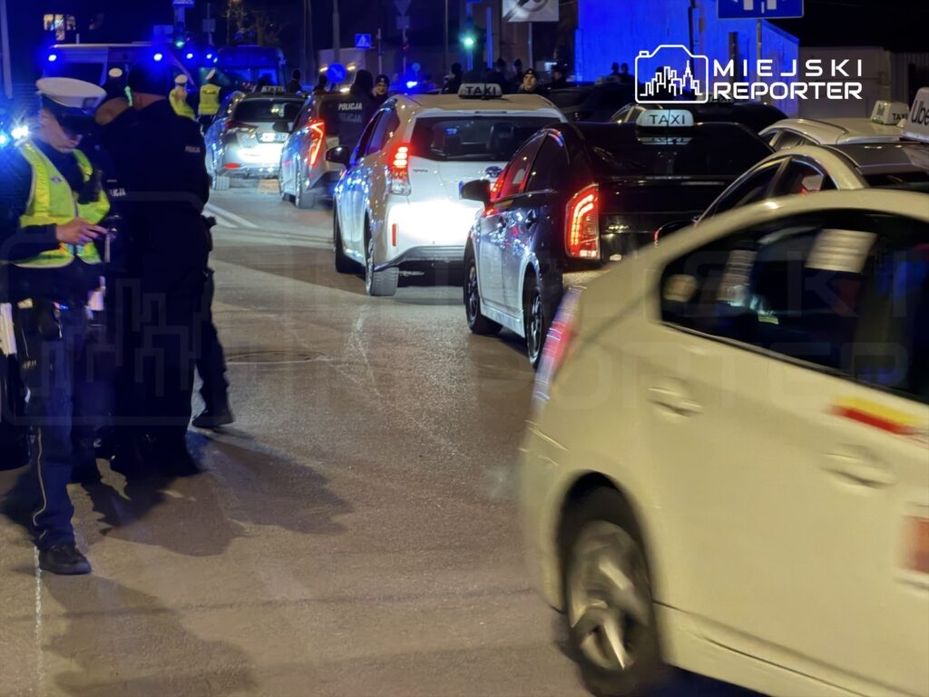 Policjanci kontrolują ruch na zatłoczonej ulicy, obok stoją taksówki z włączonymi światłami.