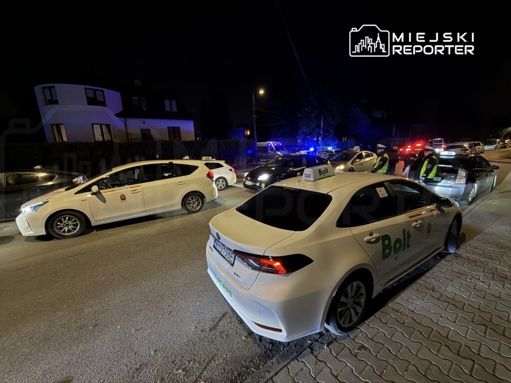 Policyjne samochody oraz taksówki stoją na ulicy w nocy, z włączonymi sygnałami świetlnymi.