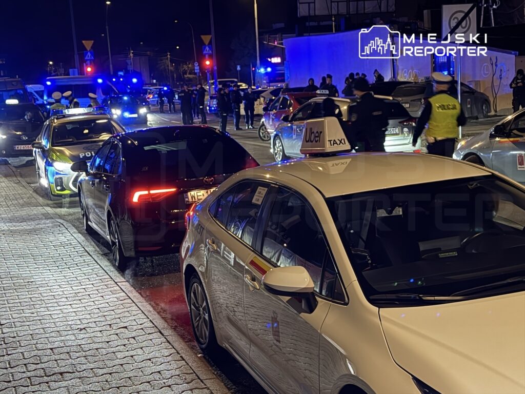 Grupa policjantów prowadzi interwencję w nocy wśród stojących samochodów na oświetlonej ulicy.