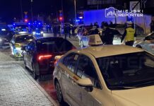 Grupa policjantów prowadzi interwencję w nocy wśród stojących samochodów na oświetlonej ulicy.