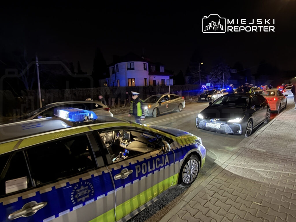 Policjant w mundurze kieruje ruchem drogowym w nocy, obok zaparkowane radiowozy i samochody osobowe.