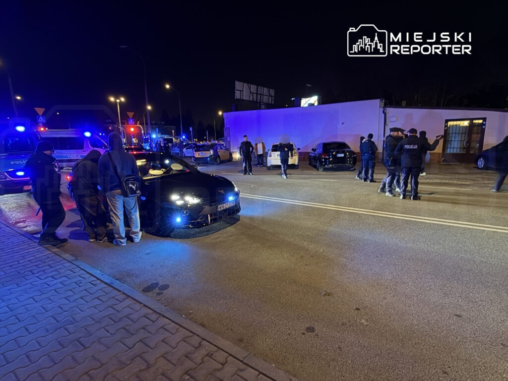 Grupa osób w ciemnych ubraniach stoi wokół czarnego samochodu sportowego, w otoczeniu radiowozów policyjnych z włączonymi sygnałami świetlnymi.