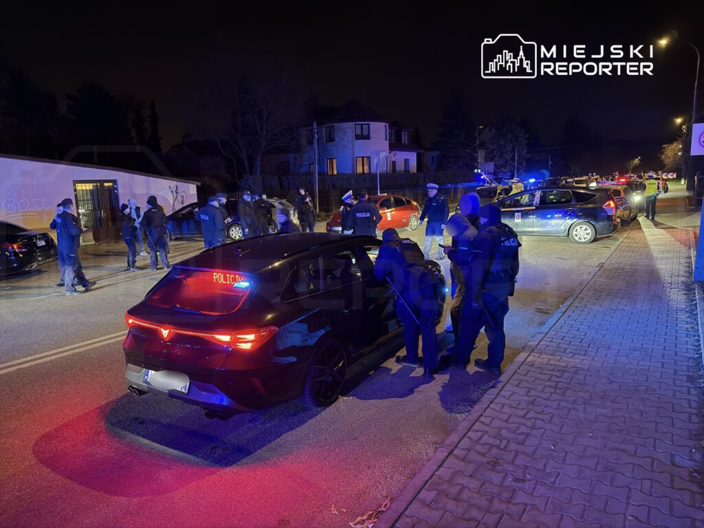 Grupa policjantów w mundurach przeszukuje samochód na oświetlonej ulicy w nocy, wokół stoją inne pojazdy.