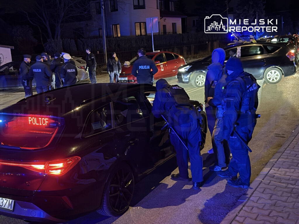 Grupa policjantów w mundurach przeszukuje samochód na oświetlonej ulicy nocą, obok stoją inne pojazdy.