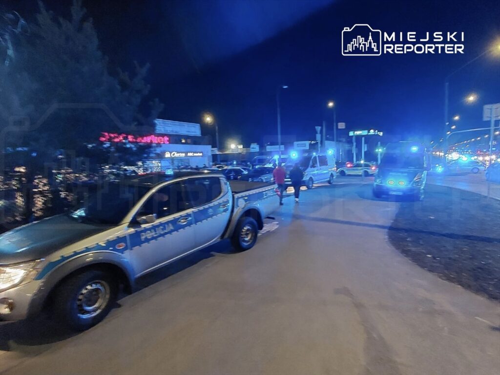 Policyjny radiowóz i karetka pogotowia z włączonymi sygnałami świetlnymi na ulicy nocą, w tle sklepy.