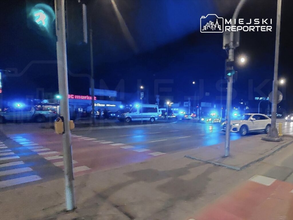 Nocna scena na ulicy z radiowozami policyjnymi zaparkowanymi w pobliżu sklepu, z migającymi niebieskimi światłami.