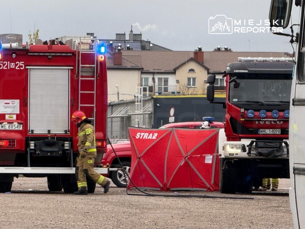 Strażak w hełmie i mundurze podchodzi do wozu strażackiego, obok rozstawiony czerwony namiot.