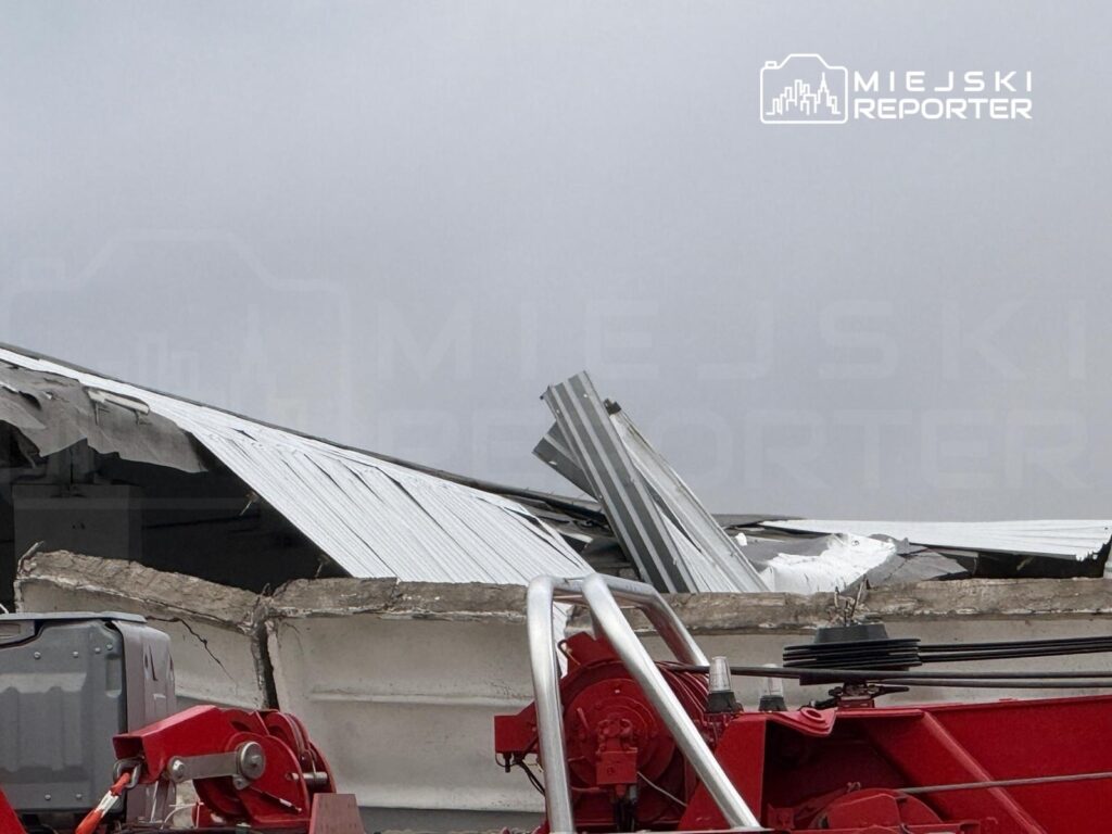 Zniszczony budynek z uszkodzoną dachówką, obok czerwony sprzęt budowlany na placu.