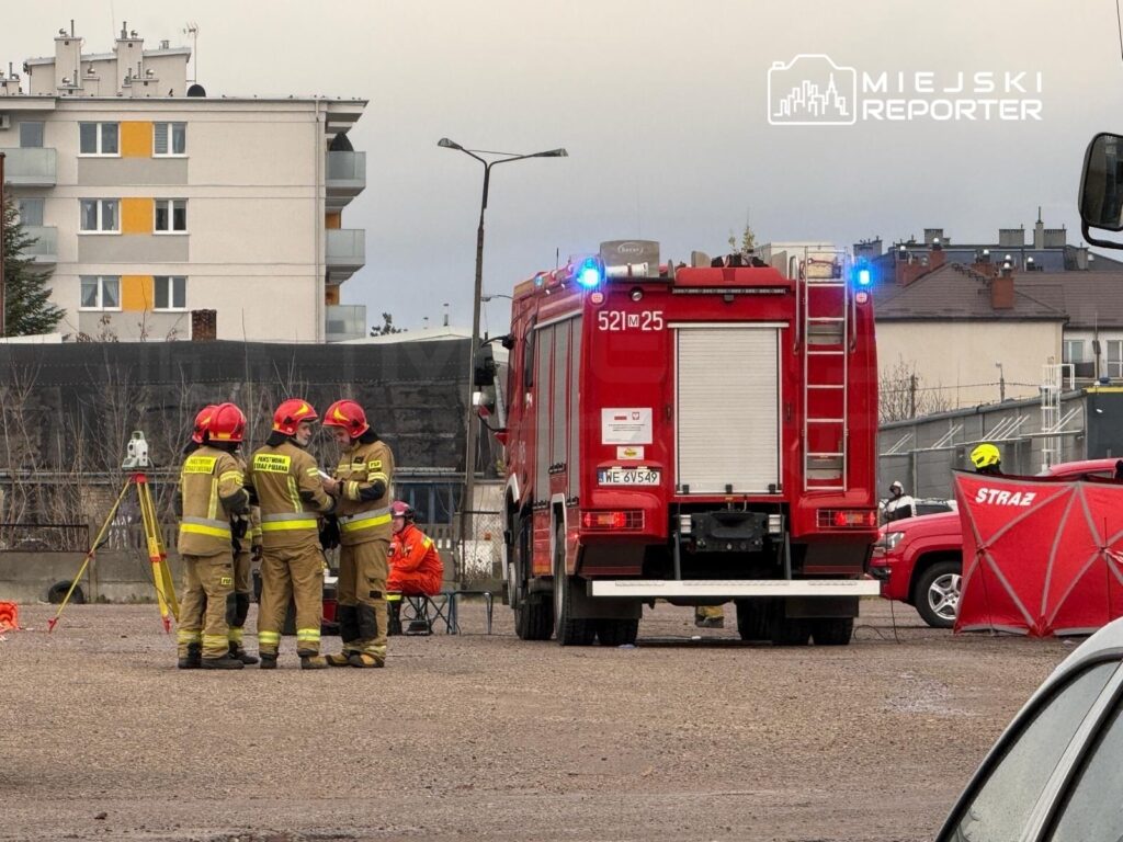 Grupa strażaków w hełmach omawia sytuację obok wozu strażackiego na placu budowy.