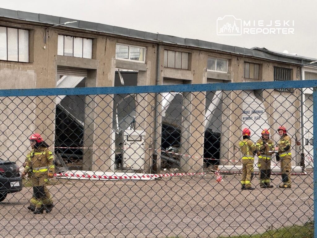 Grupa strażaków w hełmach omawia sytuację przed uszkodzonym budynkiem przemysłowym, zabezpieczonym taśmą.