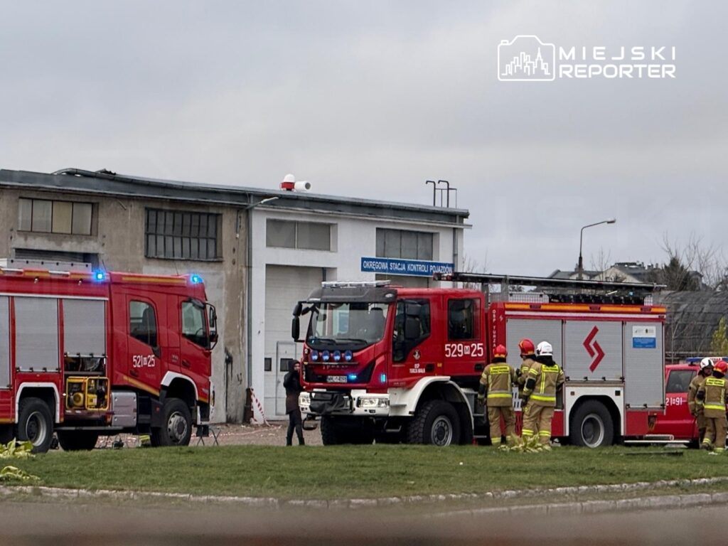 Strażacy w mundurach rozmawiają obok wozów strażackich na terenie stacji kontroli pojazdów.