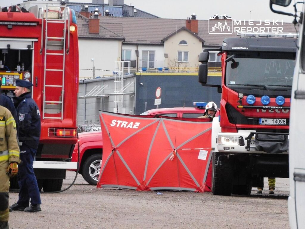 Grupa ratowników i policjantów pracuje przy czerwonym wozie strażackim, zabezpieczając teren za pomocą czerwonego namiotu.