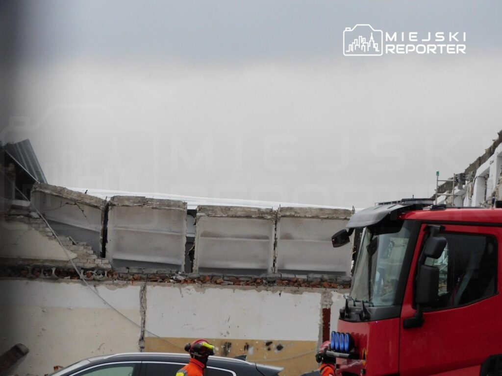 Strażak w kasku obserwuje uszkodzoną ścianę budynku, z której odpadły fragmenty, w tle widać czerwony wóz strażacki.