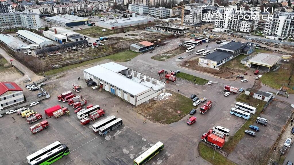 Wielka liczba wozów strażackich zaparkowanych na terenie po byłym zakładzie przemysłowym, z uszkodzonym budynkiem w tle.