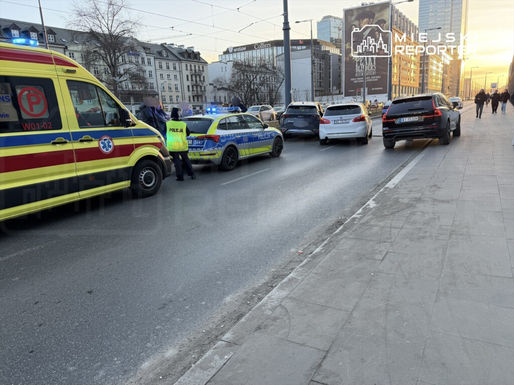 Karetka pogotowia i radiowóz policyjny zatrzymane na ulicy, obok kilku samochodów, w tle widoczne budynki i zachód słońca.