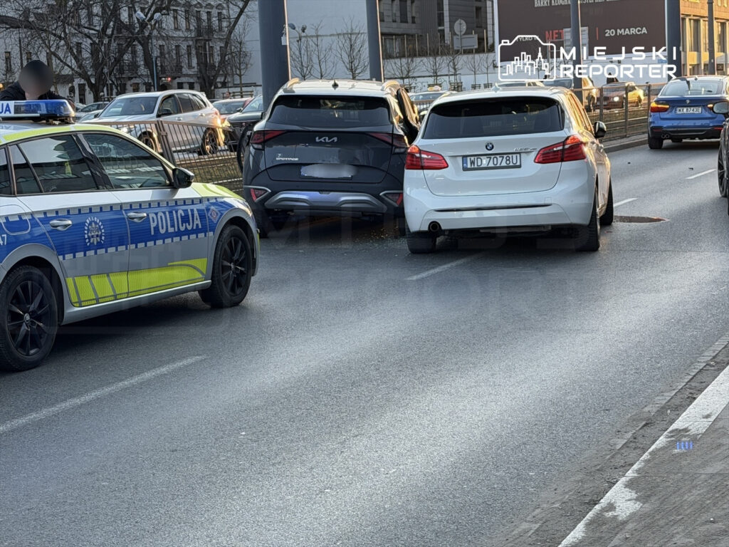 Policyjny radiowóz zatrzymany obok zderzonych samochodów na ruchliwej ulicy, w tle widoczne inne pojazdy.