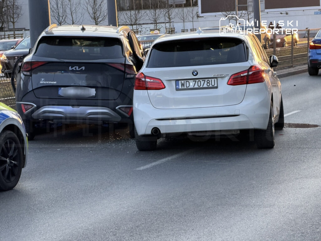 Dwa samochody osobowe, czarny SUV i biały sedan, zderzyły się na ulicy, obok widoczne inne pojazdy.