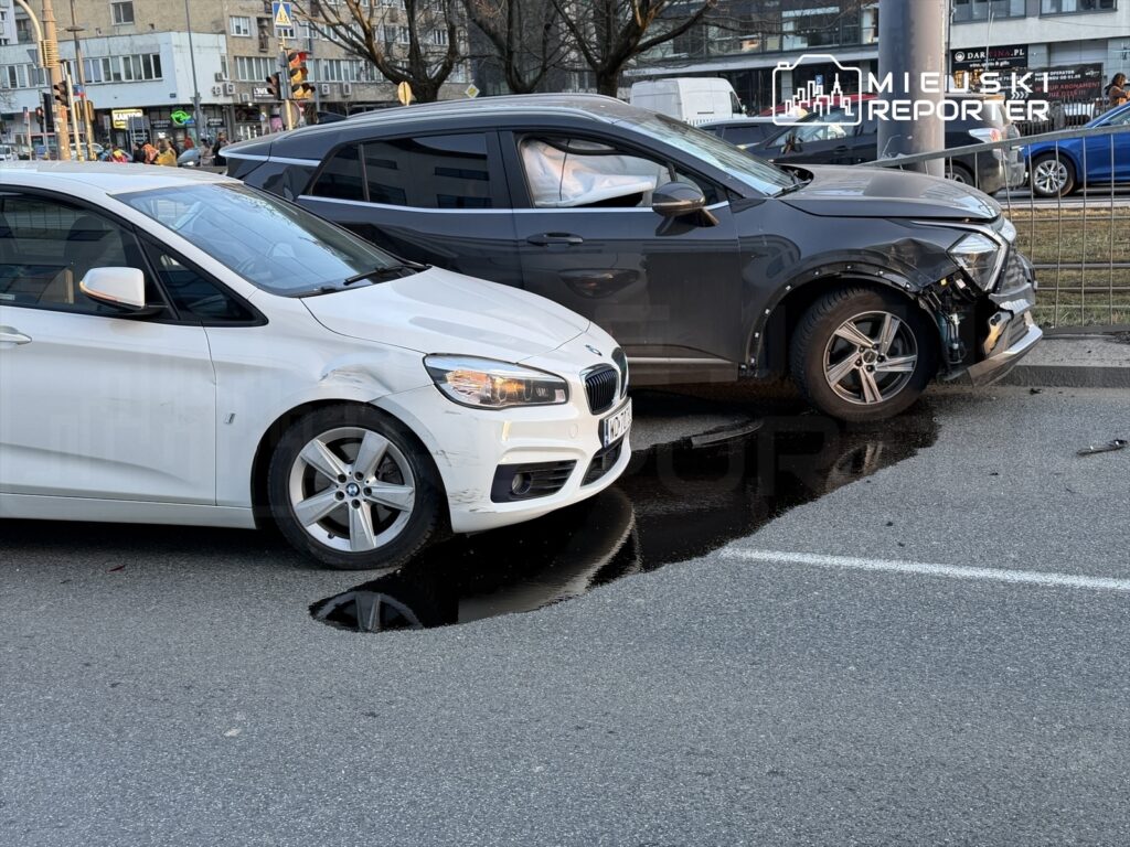 Dwa uszkodzone samochody osobowe stoją obok siebie na ulicy, z widoczną kałużą wody na jezdni.